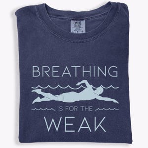 May include: Navy blue t-shirt with the text "BREATHING IS FOR THE WEAK" and a light blue swimmer graphic. The shirt is folded and has a Comfort Colors tag.