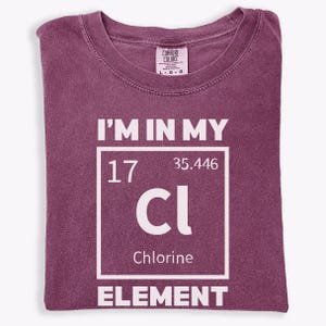 May include: A maroon t-shirt with the text "I'M IN MY" above a white box containing "17", "35.446", "Cl", and "Chlorine". The word "ELEMENT" is below the box. The shirt has a crew neck and short sleeves.