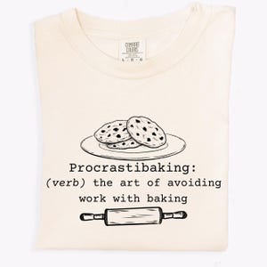 May include: A light beige t-shirt with a black and white illustration of chocolate chip cookies on a plate and a rolling pin. The text "Procrastibaking: (verb) the art of avoiding work with baking" is printed on the shirt.