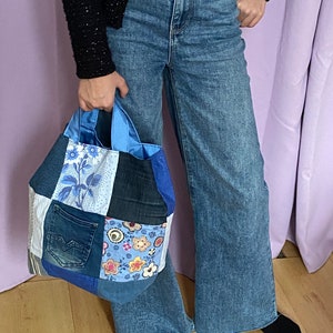 Peut inclure: Un sac fourre-tout patchwork composé de différents tons de denim bleu et de tissu à motifs. Le sac présente un mélange de motifs floraux et géométriques, avec des anses bleues et un intérieur bleu. Le sac est tenu devant une personne portant un jean bleu.