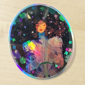May include: A holographic sticker featuring a woman with long brown hair wearing armor and holding a sword. She is surrounded by flowers and leaves. The sticker has a iridescent finish.
