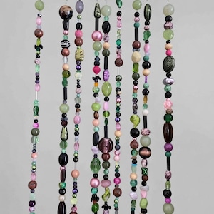 Op de afbeelding: Een verzameling kralengordijnen met diverse kleurrijke kralen in groen, roze, paars en bruin. De kralen zijn aan elkaar geregen en hangen aan een houten roede met metalen ringen.