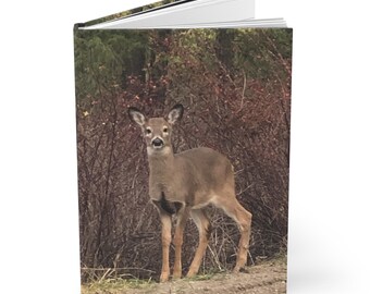 May include: A hardcover journal featuring a full-color image of a deer standing in front of a backdrop of brown and green foliage. The journal's cover wraps around the spine and back, with a clean, white interior.