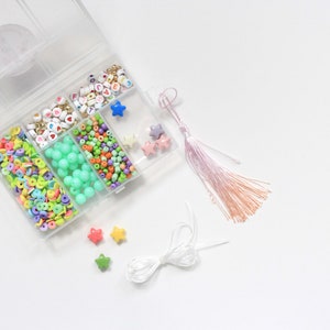 Puede incluir: Un recipiente de plástico transparente con compartimentos llenos de cuentas de colores, incluyendo letras, corazones y estrellas. También hay dos cuentas en forma de estrella, una cuerda blanca y un borla rosa.