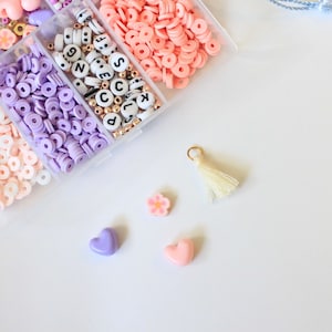 Puede incluir: Un recipiente de plástico transparente lleno de cuentas de colores, incluyendo rosa, morado y blanco. Las cuentas están dispuestas en filas e incluyen letras, corazones y flores. También hay un pequeño borla blanca.