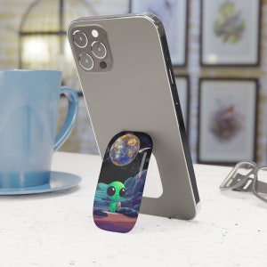 May include: A phone stand with a green alien and a planet in the background. The stand is made of a flexible material and can be used to prop up a phone.
