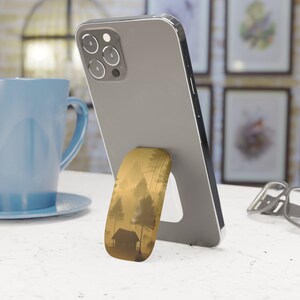 May include: A silver smartphone with a landscape-themed phone grip. The grip depicts a cabin and trees in a misty, golden scene. A blue mug and framed artwork are in the background.