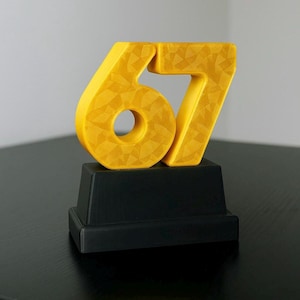 May include: A yellow number 67 sculpture with a textured surface, mounted on a black rectangular base. The sculpture is a vibrant yellow color and has a geometric pattern. The base is a solid black color.