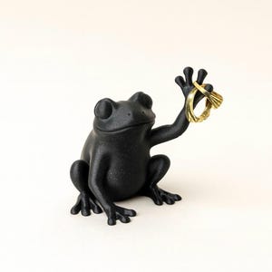以下が含まれることがあります： 2つの金の指輪を持った黒いカエルの置物。カエルは腕を上げて座り、指輪を見せています。指輪はテクスチャード加工のデザインです。置物は白い背景にあります。