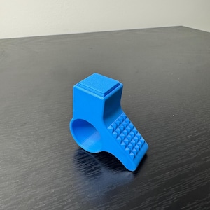 Puede incluir: Un objeto azul brillante impreso en 3D con forma de anillo. Presenta una parte superior rectangular y una superficie en ángulo texturizada con una cuadrícula de pequeños triángulos en relieve. El objeto está diseñado para usarse en un dedo.