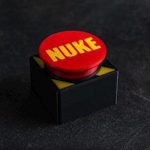 May include: A novelty button with the word "NUKE" in yellow lettering on a red circular top. The button sits on a black square base with yellow hazard stripes.