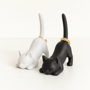 May include: Two cat-shaped ring holders, one white and one black, each with a gold ring on its tail. The cats are in a resting pose, with their heads down. The ring holders are decorative and functional.