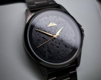 Könnte beinhalten: Eine schwarze Armbanduhr mit einem schwarzen Metallband und einem schwarzen Zifferblatt mit goldenen Akzenten. Das Zifferblatt der Uhr hat ein strukturiertes Design, das einer felsigen Oberfläche ähnelt.