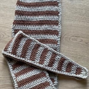 Puede incluir: Bufanda y gorro de ganchillo hechos a mano, con un diseño de rayas marrones y beige. La bufanda es larga y rectangular, mientras que el gorro es puntiagudo. Los artículos están hechos de un hilo suave y texturizado, ideal para el invierno.