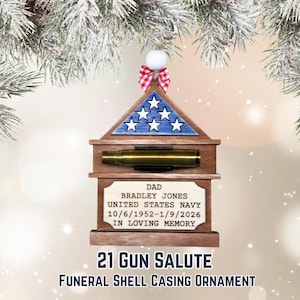 May include: A wooden ornament with a miniature American flag design and a brass shell casing. The ornament includes the text "DAD BRADLEY JONES UNITED STATES NAVY 10/6/1952-1/9/2026 IN LOVING MEMORY" and "21 GUN SALUTE FUNERAL SHELL CASING ORNAMENT".
