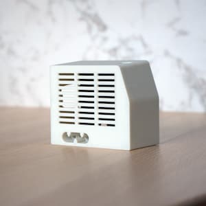 Op de afbeelding: Een witte plastic doos met een rechthoekige ventilatieopening en een kleine opening aan de voorkant. De doos heeft een gladde, matte afwerking.
