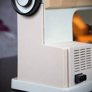 May include: A white sewing machine with a black knob and a beige top. The machine has a built-in power cord and a vent on the side.