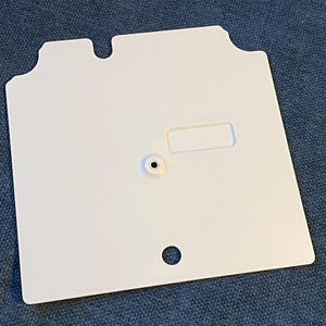 May include: A flat, square white plastic component featuring a central circular hole, a small rectangular indentation, and a distinct notched top edge with two protruding tabs. The surface is smooth and uniform, designed for mechanical assembly.