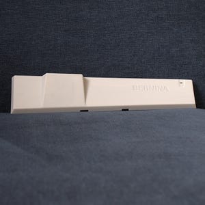 Op de afbeelding: Een lichtbeige naaimachine-accessoire met de merknaam "BERNINA" erop gedrukt. Het accessoire heeft een rechthoekige vorm met een trapsgewijs ontwerp en verschillende kleine zwarte openingen. Het ligt op een donkerblauwe ondergrond.