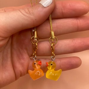 Puede incluir: Dos encantos de patito de goma amarillo con picos naranjas y eslabones de cadena dorados. Los encantos están unidos a cordones dorados.