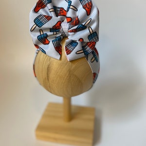 May include: A white hair bow with a red, white, and blue popsicle print. The bow is displayed on a wooden stand.