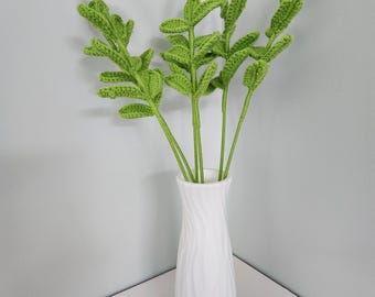 May include: A white ceramic vase with a textured, wavy design holds several stems of crocheted green leaves. The leaves are a vibrant green, and the vase is set against a neutral background, creating a simple, elegant display.