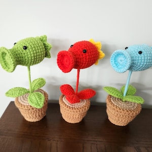 Puede incluir: Tres plantas de ganchillo hechas a mano en macetas de terracota. Las plantas son verdes, rojas y azules, cada una con un diseño único. La planta verde tiene una boca grande y abierta, la planta roja tiene un acento amarillo y la planta azul tiene una boca ancha y abierta.