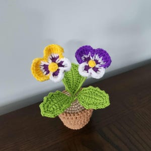 May include: A crocheted potted plant with two flowers. One flower is yellow and purple, the other is purple and white. The pot is brown.