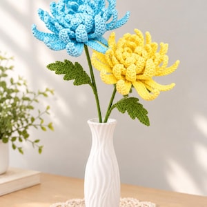 Pode incluir: Duas flores de crochê, uma azul-clara e uma amarela, com caules e folhas verdes, dispostas num vaso de cerâmica branco, alto e esguio, com uma textura ondulada vertical. As flores apresentam pétalas em camadas feitas de fio.