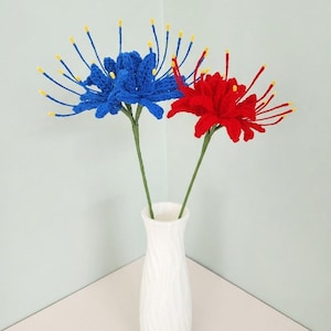 May include: Two crocheted spider lily flowers, one in vibrant blue and one in bright red, each featuring long, thin petals with yellow tips. The flowers are attached to green stems and are displayed together in a tall, white, textured vase.