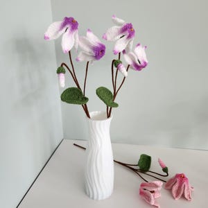 May include: A white, wavy-textured vase holds several crocheted cyclamen flowers with white and purple petals, green leaves, and brown stems. Two additional crocheted flowers lie on the surface, featuring pink and white petals with green buds.