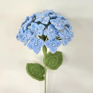 May include: A crocheted blue hydrangea flower with green leaves and white pearl accents.