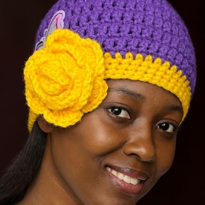 May include: A handmade crochet hat in purple with a yellow brim and a large yellow flower. A small butterfly applique is visible on the side of the hat. The hat is a vibrant accessory.