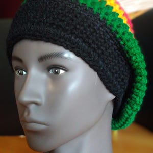 May include: A black, green, yellow, and red crocheted beanie hat. The hat is displayed on a mannequin head. The hat has a black base with green, yellow, and red stripes at the top. The hat is made of a thick, textured yarn.