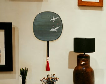 Peut inclure: Éventail rond bleu canard avec manche noir et pompon rouge. Il présente deux grues blanches en vol. L'éventail est accroché à un mur blanc, avec une lampe et d'autres objets décoratifs en arrière-plan.