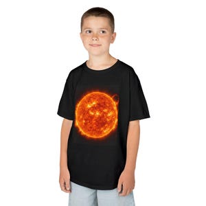 Peut inclure: Un t-shirt noir à manches courtes avec une grande impression graphique détaillée du soleil sur la poitrine. Le motif solaire présente des teintes vibrantes d'orange, de jaune et de rouge avec des éruptions solaires et une texture visible.