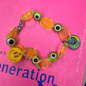 May include: A yellow and green plastic bracelet with smiley face, eye, and "Shell Bead" charms.