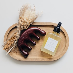 Könnte beinhalten: Ein ovales Holztablett enthält ein dunkelbraunes Kopfhautmassagegerät aus Holz, eine quadratische Glasflasche mit botanischem Kopfhautöl mit schwarzem Tropfverschluss und ein kleines Bündel getrockneter beiger Blumen, die mit Schnur gebunden sind.
