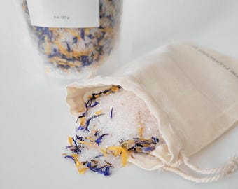 Puede incluir: Una bolsa transparente y una pequeña bolsa con cordón llena de sales de baño. Las sales se mezclan con pétalos de flores amarillas y azules secas. La bolsa del fondo tiene una etiqueta que dice "EPSOM SALTS - HIMALAYAN PINK SALT".