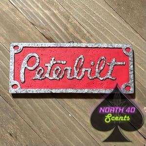 May include: A rectangular red sign with the word "Peterbilt" in silver script. The sign has a silver border and is mounted on a wooden surface. The North 40 Scents logo is in the lower right corner.