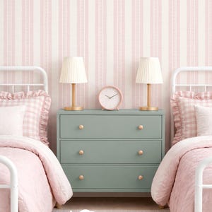 May include: A sage green three-drawer dresser with round wooden knobs, flanked by two white metal beds with pink gingham bedding. Two small lamps with pleated shades and a round pink clock sit on the dresser against pink striped wallpaper.
