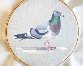 May include: Cross-stitch embroidery of a pigeon in a wooden hoop. The bird is depicted in shades of gray, purple, and green, with pink feet and a black beak. The artwork is on a light gray fabric.