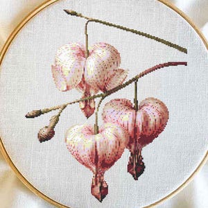 Può includere: Un ricamo a punto croce di fiori di cuore sanguinante rosa con steli e foglie verdi su uno sfondo bianco.