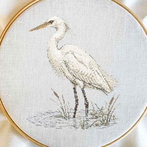 May include: A circular wooden embroidery hoop containing a cross-stitch pattern of a white egret standing in shallow water with reeds. The bird is rendered in shades of white and gray, while the water and reeds feature muted brown and green tones.