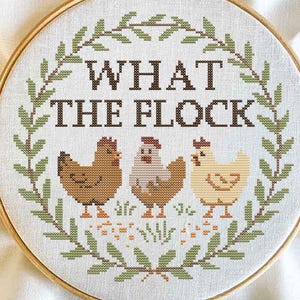 May include: Cross-stitch embroidery in a wooden hoop features the phrase "WHAT THE FLOCK" above three chickens. The chickens are brown, beige, and yellow, surrounded by a green leaf border. A charming, rustic home decor item.