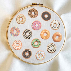Könnte beinhalten: Nahaufnahme eines runden Stickrahmens mit weißem Stoffhintergrund und 14 bunten Donuts, die in einem Kreis gestickt sind.
