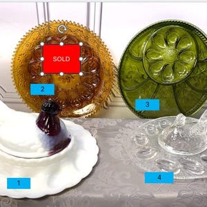 May include: A collection of vintage serving dishes. Includes a white hen-shaped dish with a purple and white accent, a golden-yellow plate, a green plate, and a clear glass egg serving tray. The golden plate is marked as "SOLD".
