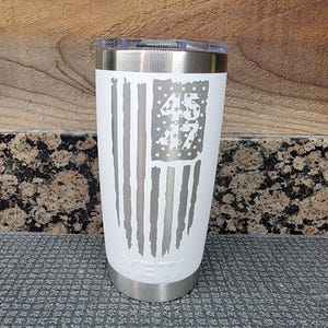Pode incluir: Um copo isolado branco com aro e tampa prateados. O design apresenta uma bandeira americana desgastada com os n&uacute;meros "45" e "47" no campo de estrelas. A marca "YETI" &eacute; vis&iacute;vel na parte inferior.