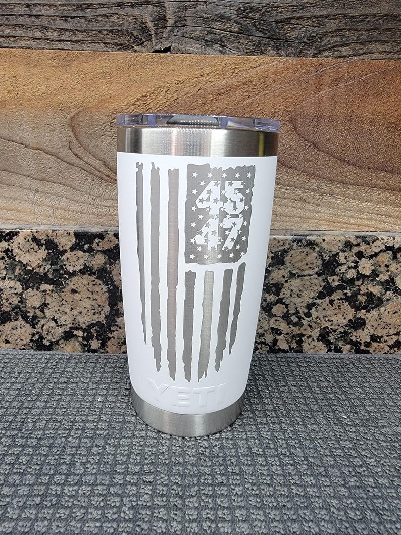 Pode incluir: Um copo isolado branco com aro e tampa prateados. O design apresenta uma bandeira americana desgastada com os n&uacute;meros "45" e "47" no campo de estrelas. A marca "YETI" &eacute; vis&iacute;vel na parte inferior.