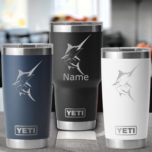 May include: Three insulated tumblers in navy blue, black, and white, each featuring a marlin fish design and the brand name "YETI." The black tumbler has the word "Name" below the fish. The tumblers have silver rims.
